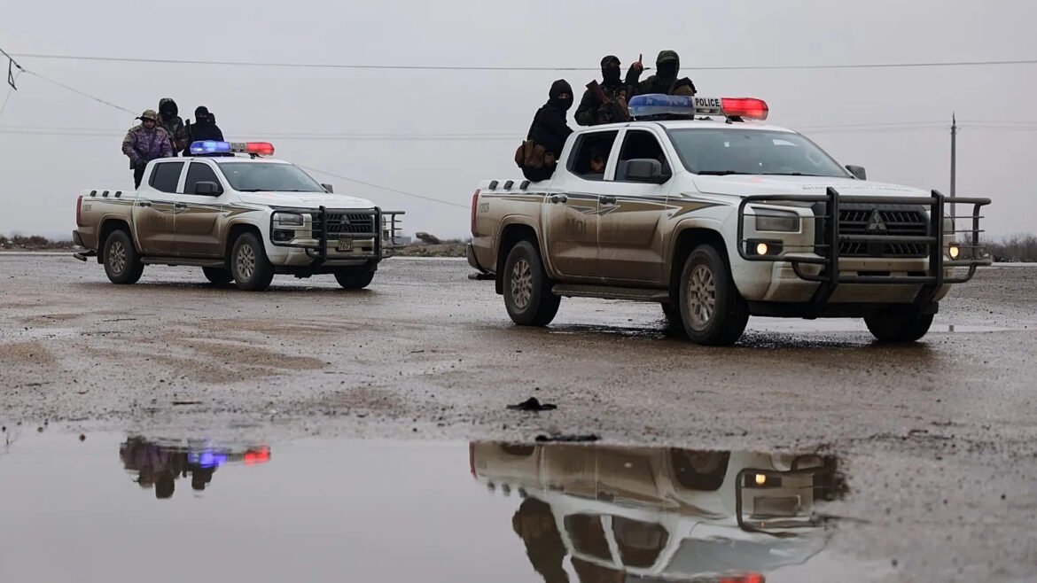 Armed police officers ride in two muddy pickup trucks with red-blue lights on a rain-soaked road; reflected in a large puddle below.