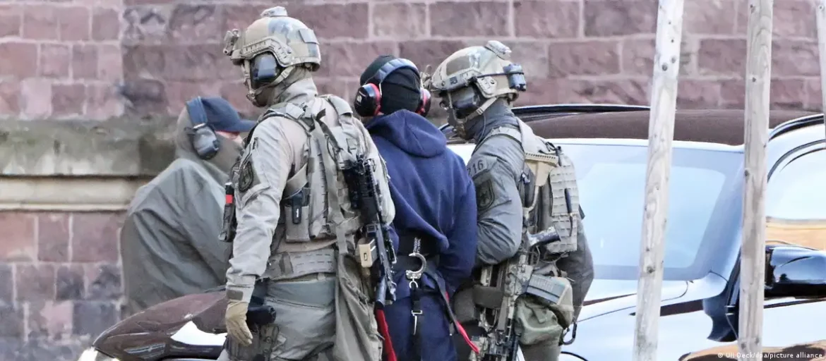 Tactical officers in body armor escort a detainee in a blue hoodie beside a car.