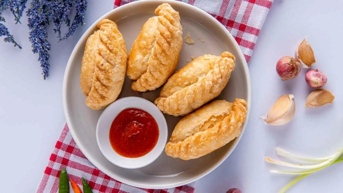 Plate of four golden baked pastries with a red dipping sauce in a small bowl on a light gray surface, with lavender, garlic, and a red-checkered cloth nearby.