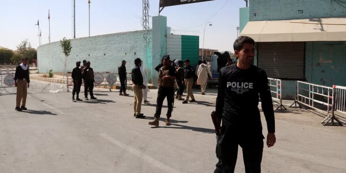 Police officer in black uniform stands at a barricaded street, with several civilians and other officers gathered near a turquoise building in the background.
