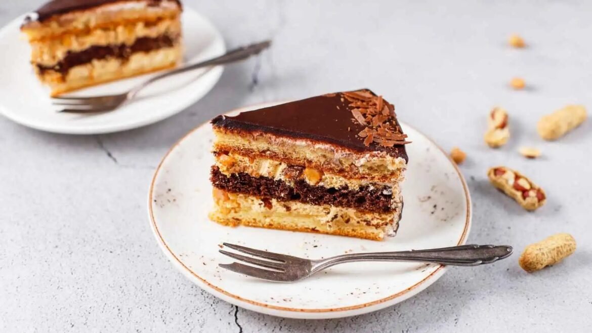 Slice of multi-layer chocolate cake with cream filling and glossy ganache on a white plate, fork beside it.