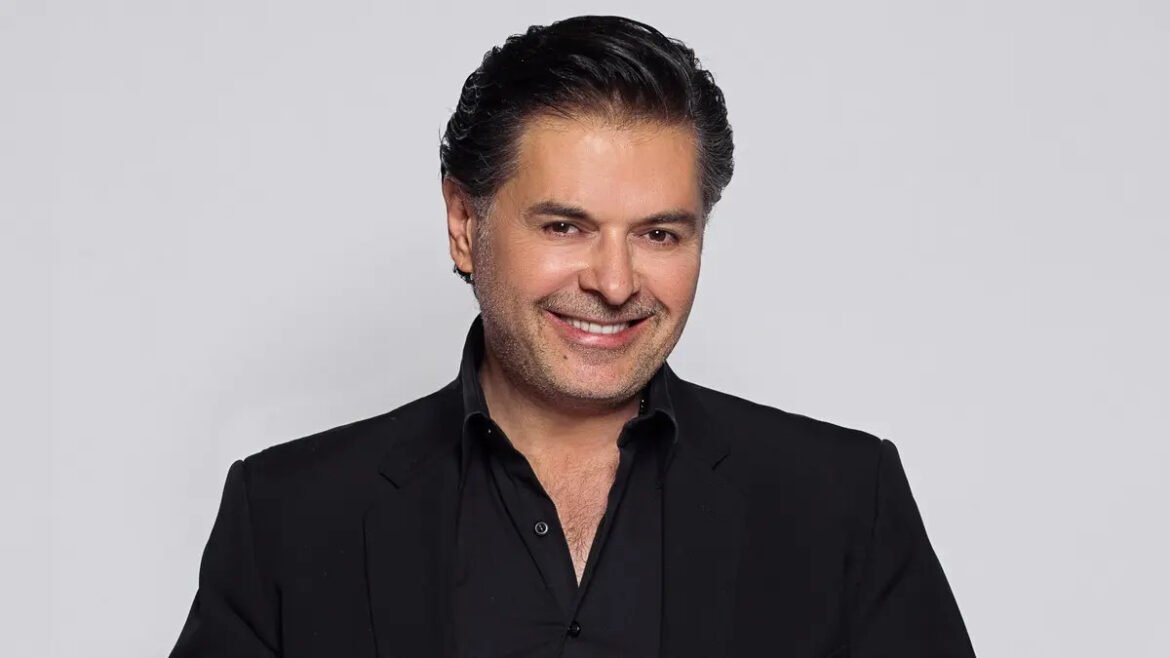 Head-and-shoulders portrait of a smiling man in a black blazer against a gray background, studio photo of a professional-looking man.