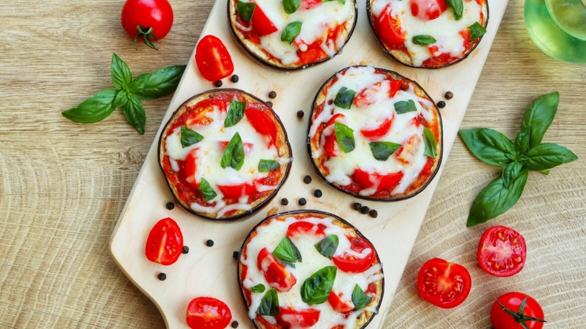Eggplant pizzas on a wooden board topped with mozzarella, tomato, and basil leaves, surrounded by cherry tomatoes and fresh basil.