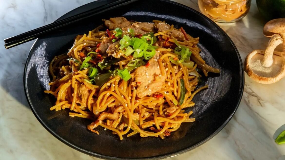 Plate of sesame beef and vegetable stir-fry noodles topped with chopped green onions on a black plate with chopsticks nearby, served on a marble surface, close-up