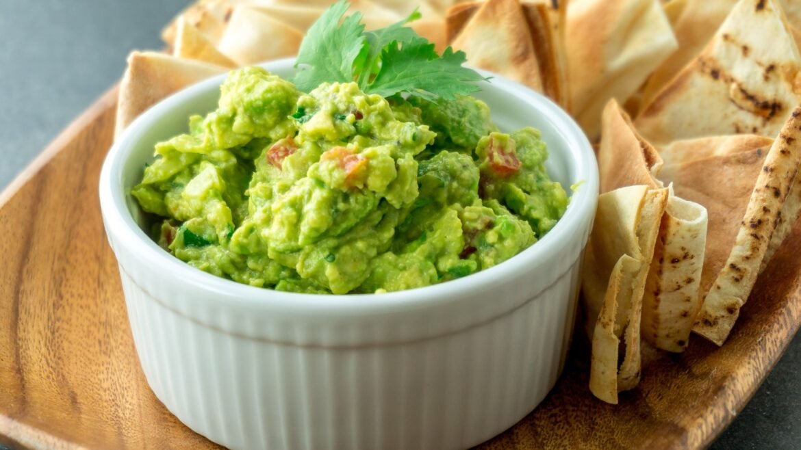 White ramekin of chunky guacamole with tomato bits and a cilantro garnish, flanked by warm tortilla chips on a wooden tray