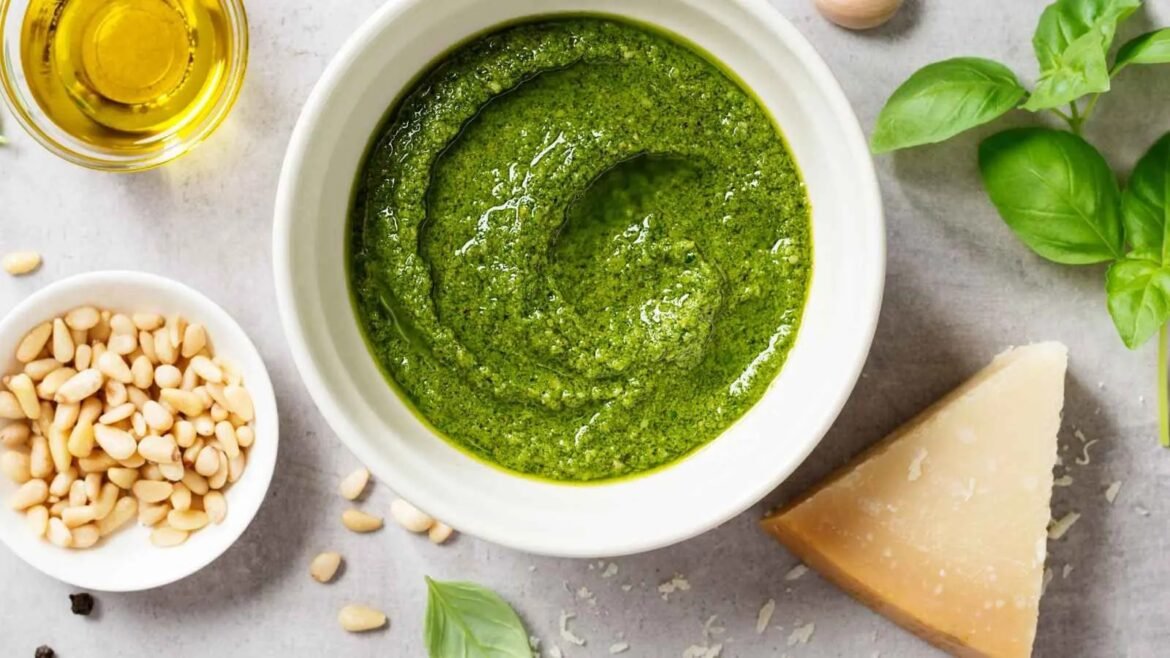 White bowl holds bright green pesto surrounded by pine nuts, olive oil, basil leaves, and a Parmesan wedge.