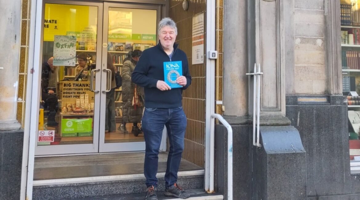 Smiling man in a dark sweater stands in the doorway of a bookshop, holding a blue-covered book toward the camera.