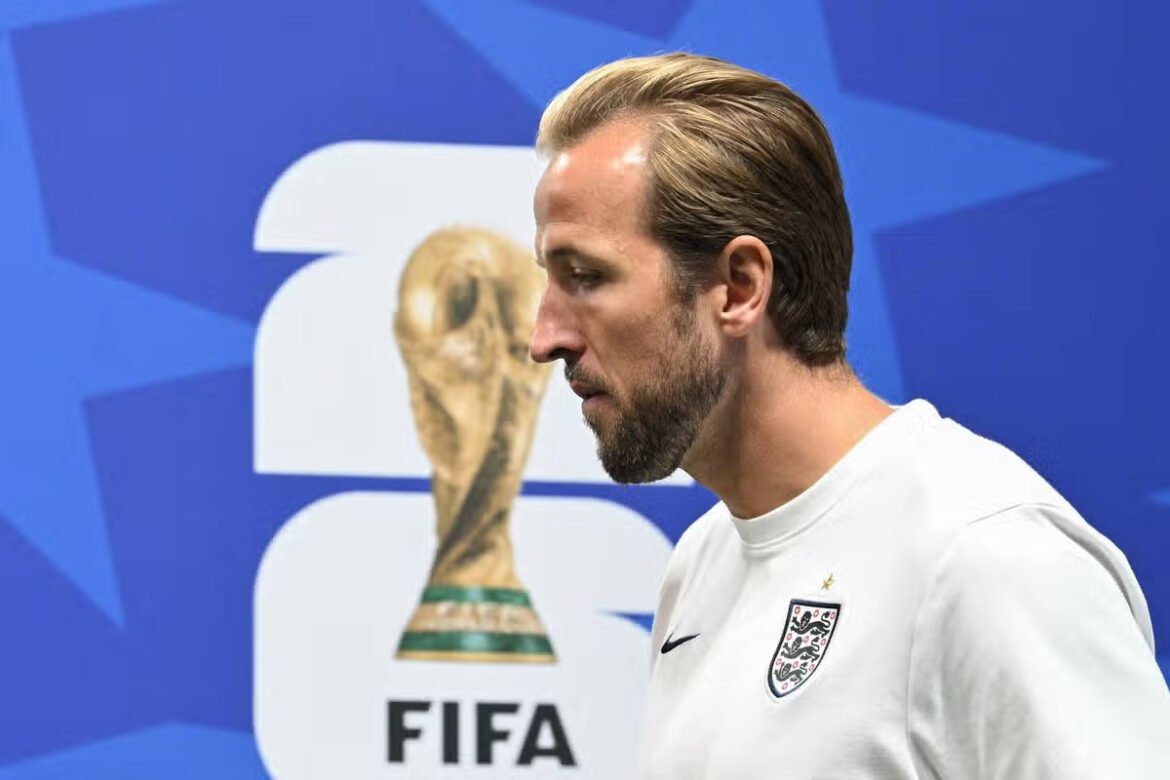 Man in a white England football shirt faces left as a large FIFA World Cup trophy graphic looms behind him on a blue background