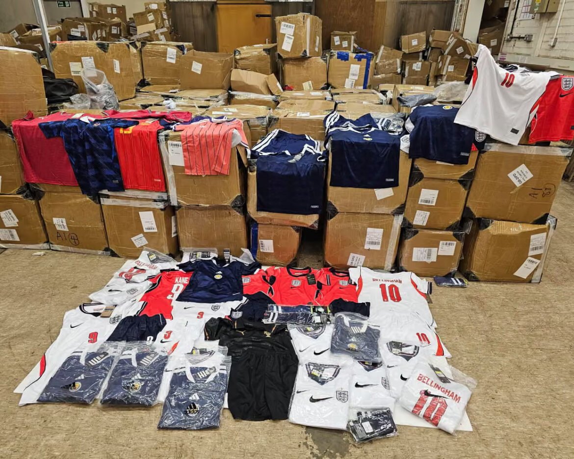 Warehouse scene with stacked cardboard boxes and a pile of England rugby/football jerseys laid out on the floor for sorting or display.