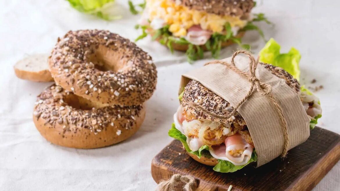 Seeded bagels stacked on a light surface beside a wrapped sandwich with lettuce and deli meat on a wooden board.