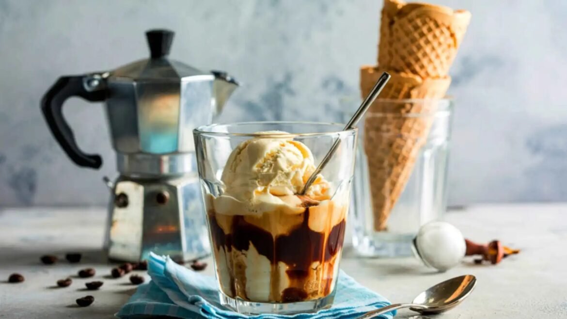 Clear glass cup with vanilla ice cream and chocolate sauce drizzle, sitting on a blue napkin; espresso pot and waffle cones in the background tarjoaa dessert scene