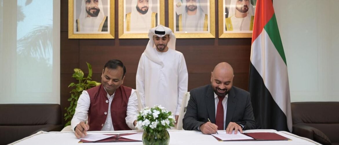 Two men signing documents at a formal table, with a UAE flag and framed portraits behind them.