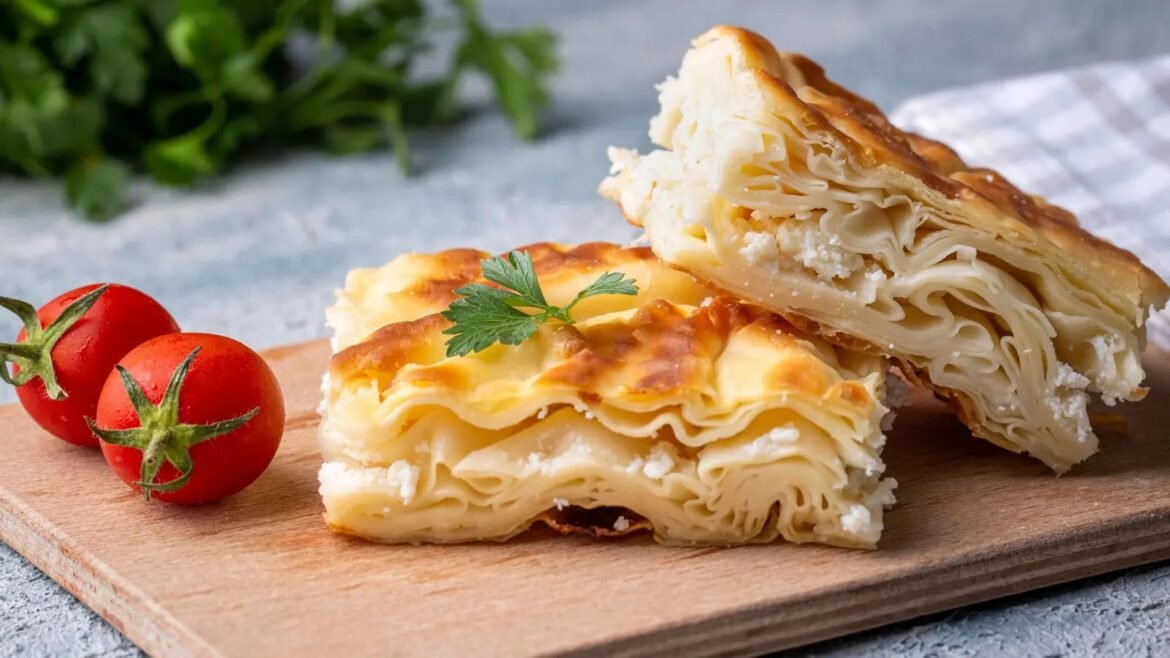 Two ripe tomatoes beside a stack of golden, flaky cheese-filled phyllo pastries on a wooden board, with a parsley garnish on top.