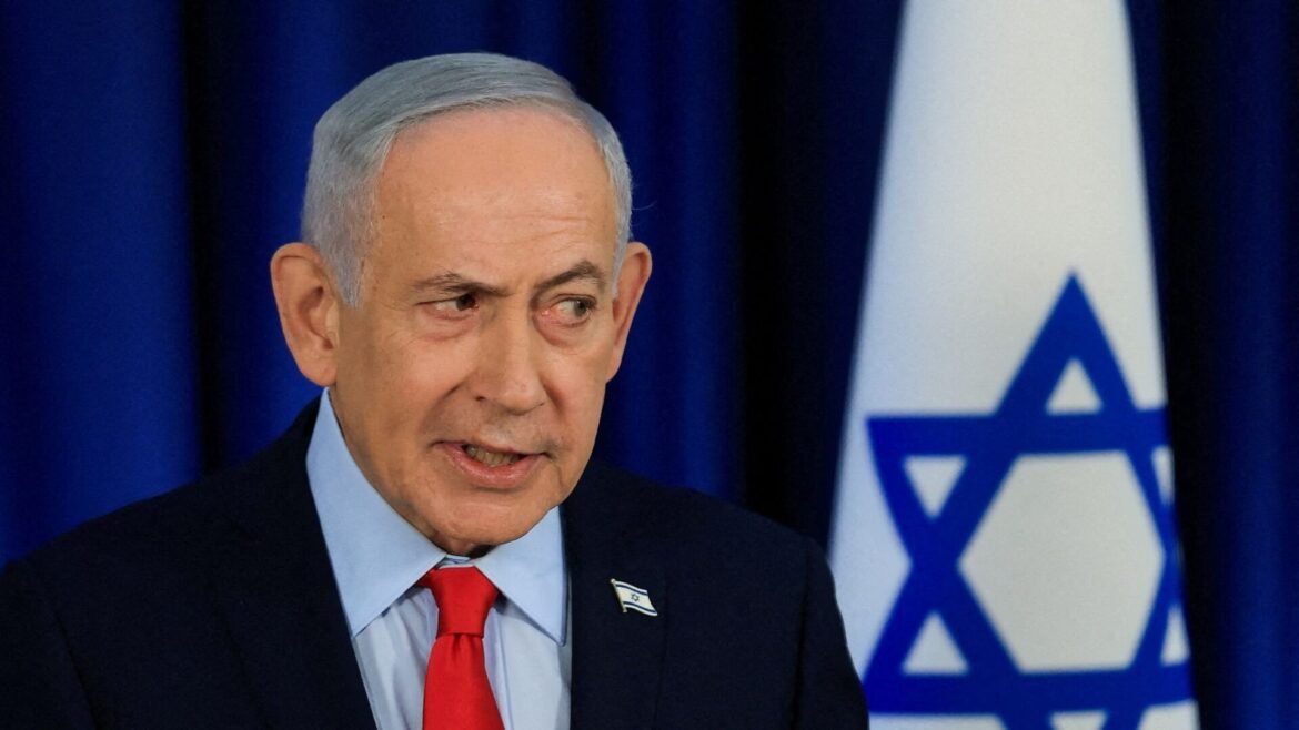 Man in a dark suit and red tie speaks in front of an Israeli flag backdrop, with a blue curtain behind.
