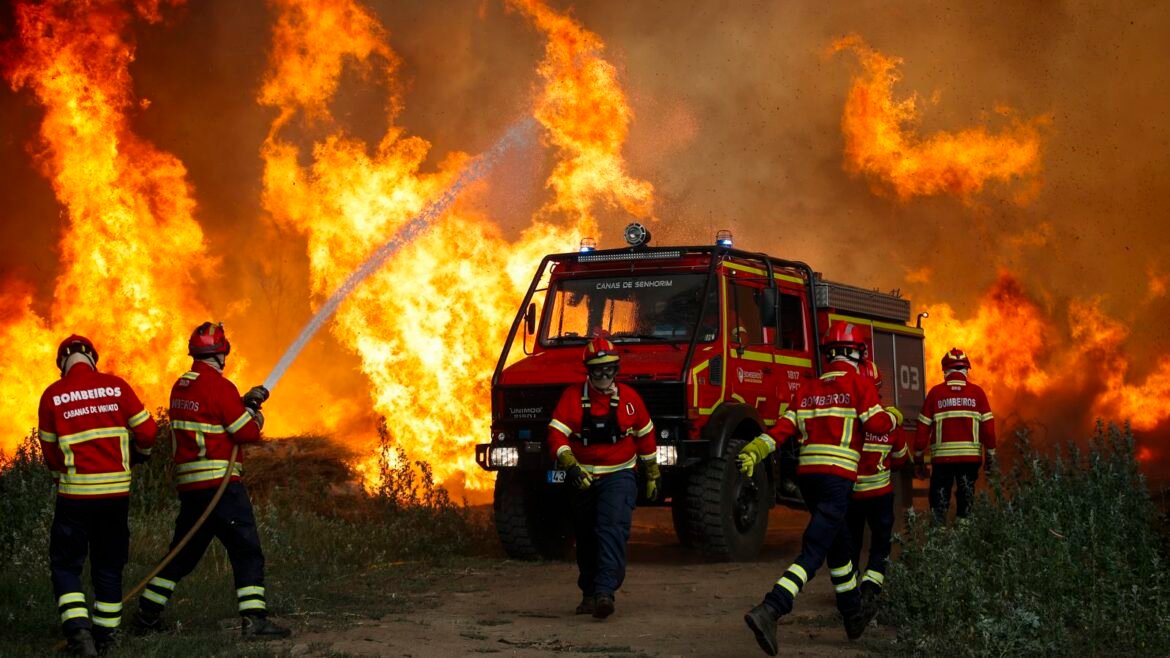 Firefighters in red gear spraying water at a large wildfire with a red truck nearby, flames engulfing the area.
