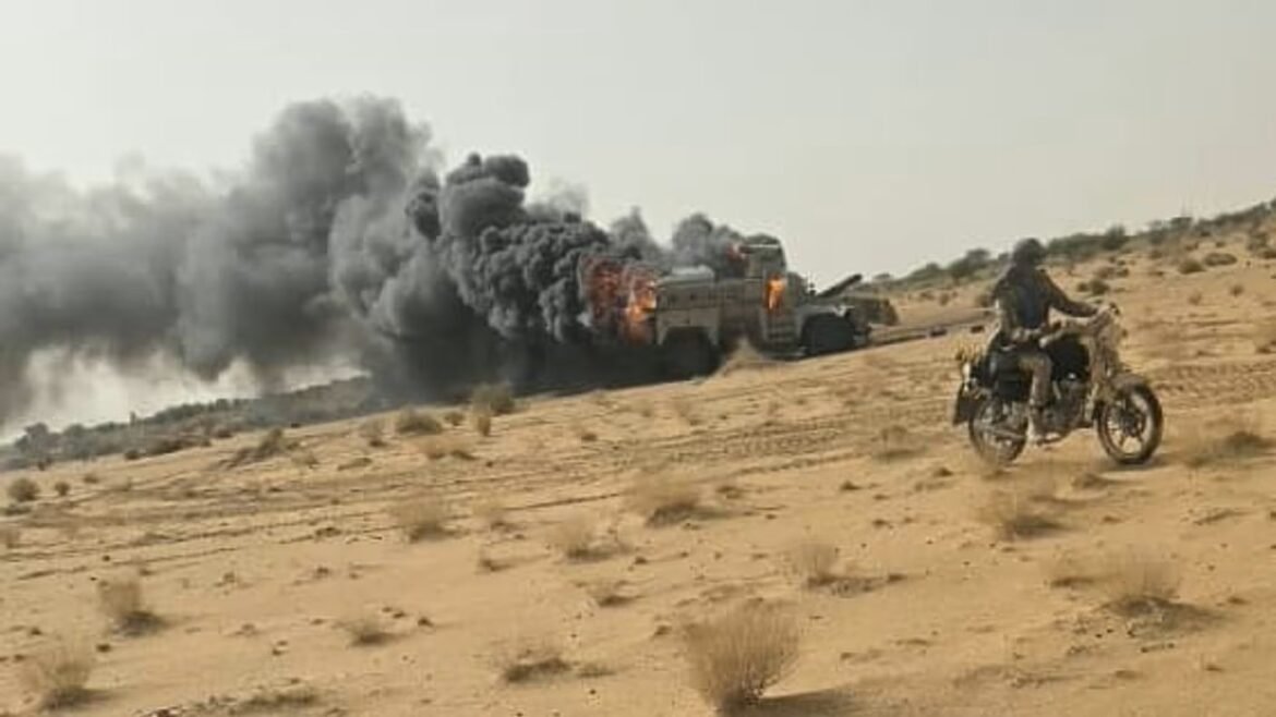 Desert scene with a burning military truck and thick black smoke, a motorcyclist riding nearby in the foreground.