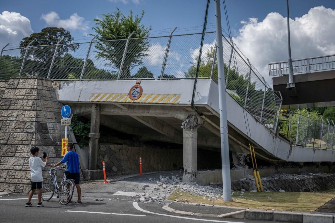 Under a collapsed overpass with debris on the ground; two boys with a bicycle stand nearby, examining the scene as a pedestrian/bike path runs beneath a fenced bridge.