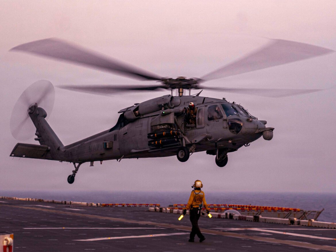 US Navy helicopter hovering above the deck of an aircraft carrier, crewman in orange flight suit nearby as rotor blades spin.