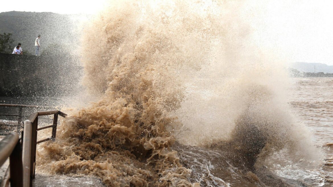 A large muddy wave crashes over a railing at a waterfront, spraying water toward onlookers standing on a concrete jetty edge to the left.