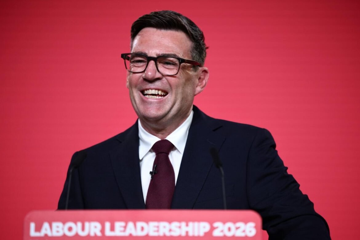 Man in a dark suit and maroon tie smiles while giving a speech at a Labour Leadership 2026 event, red backdrop.