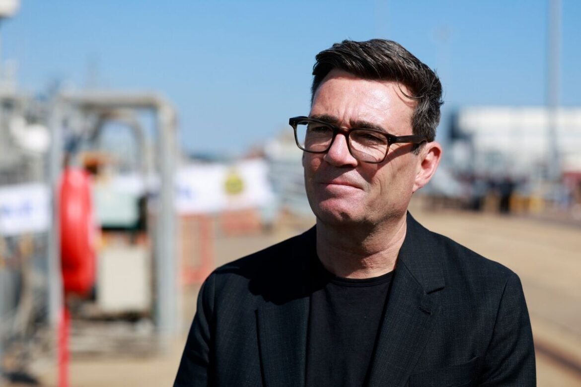 Man in a black blazer and glasses outdoors, looking thoughtful near an industrial area with equipment in the background.