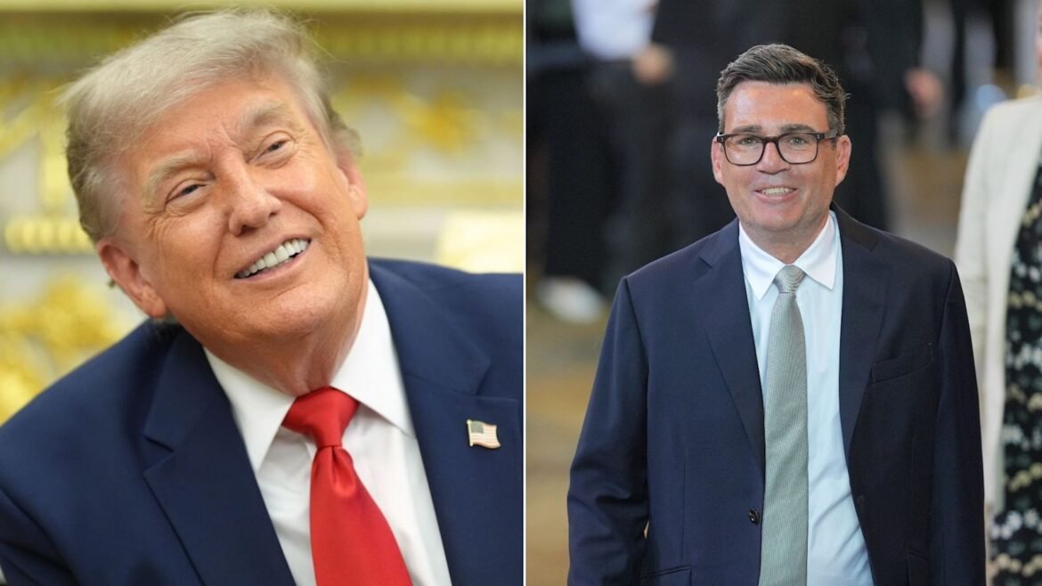 Two men in navy suits at a formal setting: left man with light hair and red tie smiling, right man with glasses and gray tie.