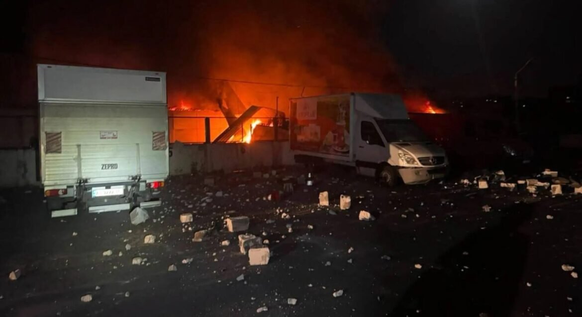 Nighttime industrial fire with orange flames; white delivery trucks parked near a burning building, debris scattered on the ground.