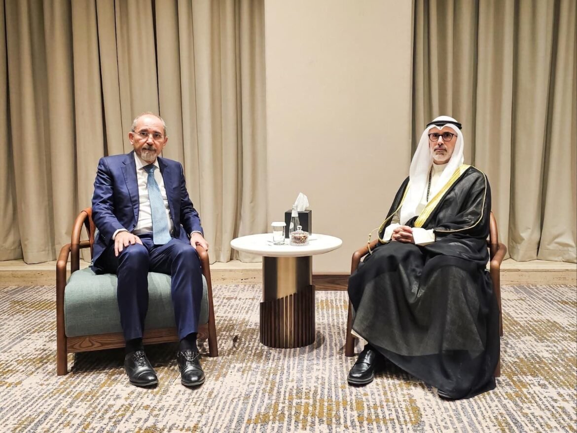 Two men sit facing each other in a formal meeting: one in a blue suit, the other in traditional Arab attire with a white headscarf and black robe, separated by a small table with water and tissues.