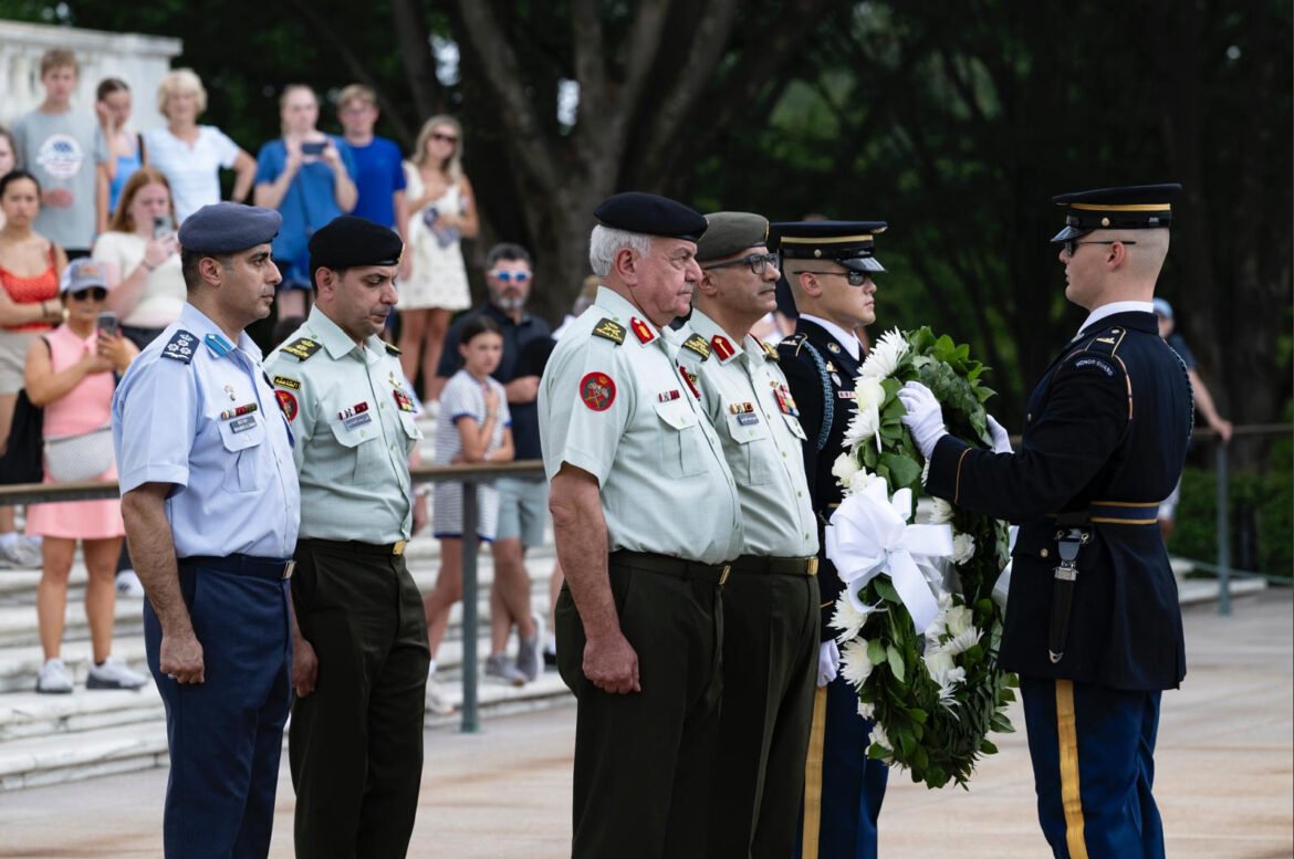 Military officers stand in a line as a uniformed guard presents a white wreath to them during a ceremony, with onlookers in the background.