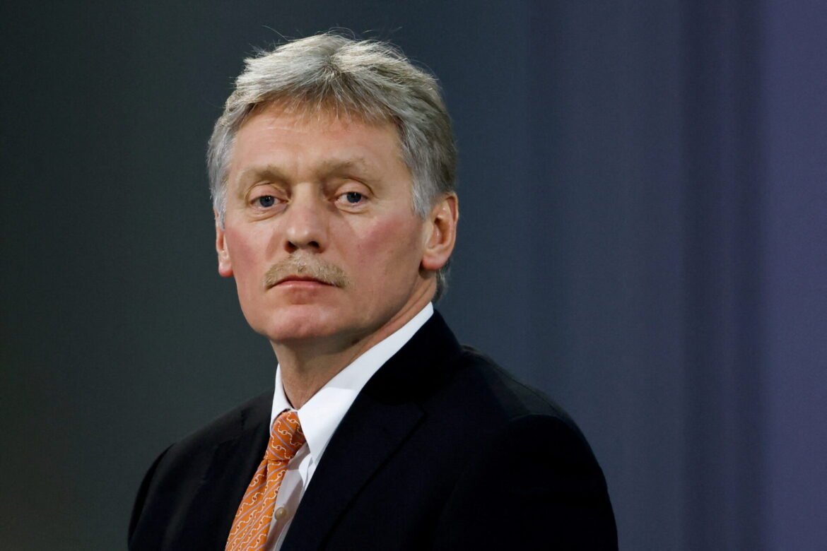Portrait of a man in a dark suit with an orange patterned tie, looking to the left against a dark backdrop.