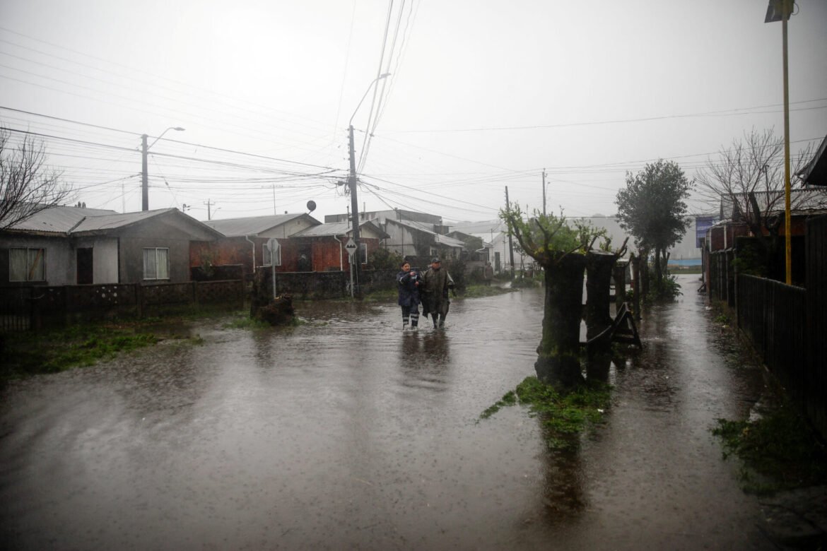 Flooded suburban street with two people in rain gear walking through ankle-deep water among submerged houses and tangled power lines overhead.