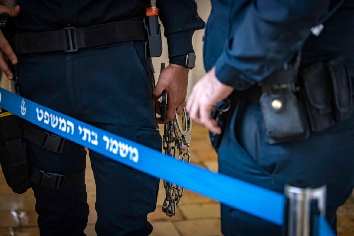 Two police officers stand behind a blue barrier tape, one holding handcuffs and chains while wearing duty gear and a smartwatch.