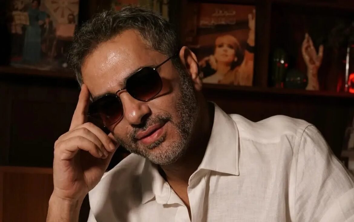 Man with salt-and-pepper hair and beard, wearing sunglasses and a white shirt, resting his head on his hand in a dimly lit lounge or bar.