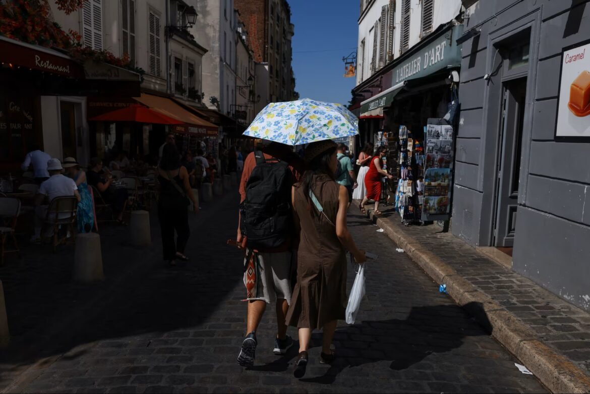 Two women walk away down a sunlit cobblestone street, sharing a blue floral umbrella, with cafes on the left and shops on the right nearby.