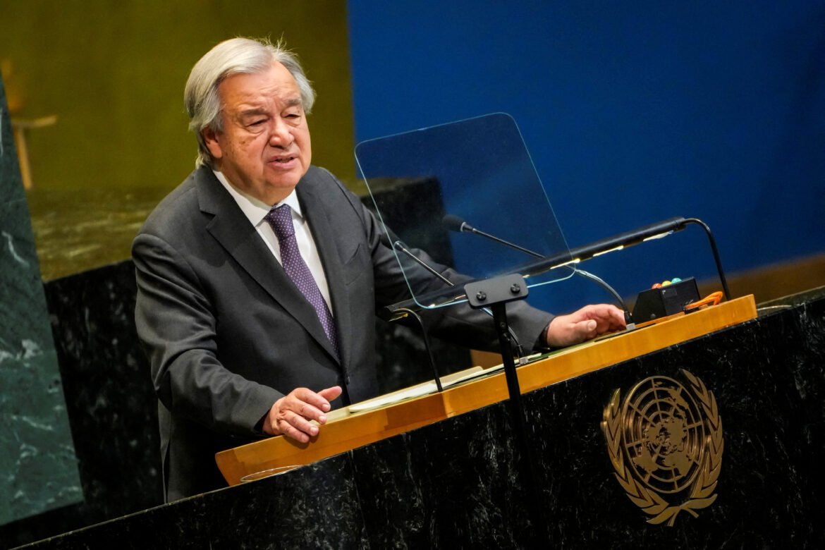 Older man in a dark suit and purple tie speaks at a United Nations podium with a clear protective panel in front, blue backdrop behind him.