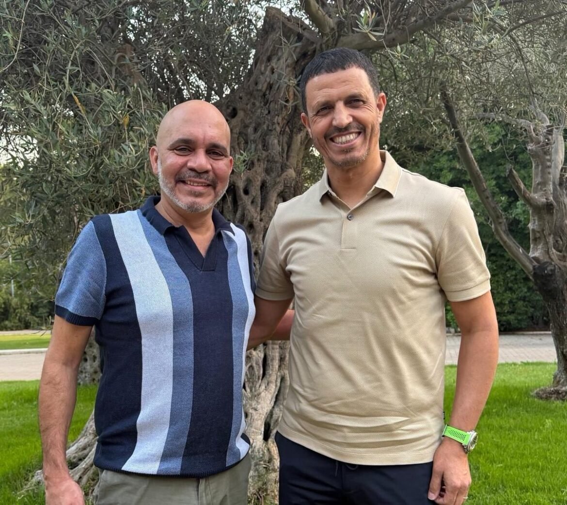 Two men standing outdoors beside a tree, smiling at the camera.