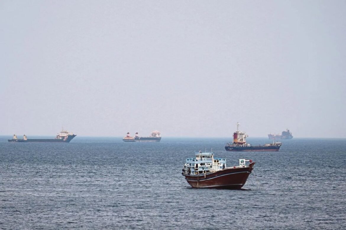 A brown passenger boat in the foreground on a calm blue sea with several cargo ships in the hazy distance.
