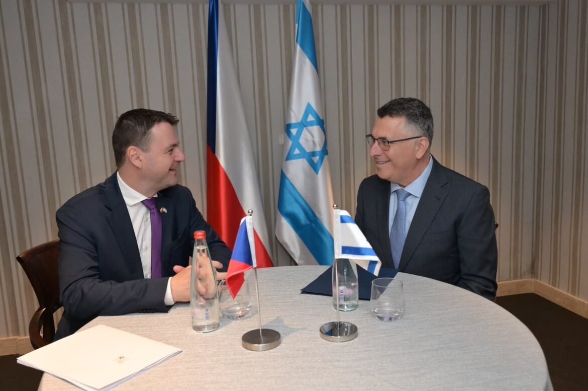 Two businessmen sit at a round table, smiling during a discussion, with Czech, Israeli, and Greek flags on display between them.