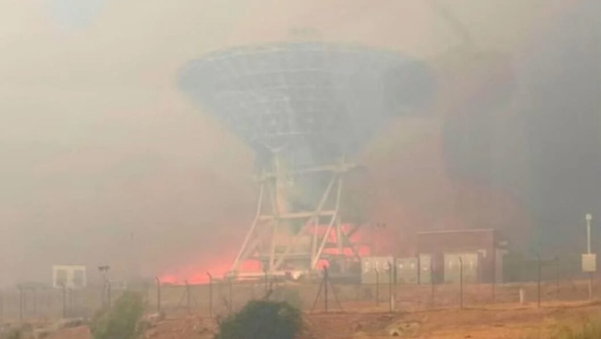 Large satellite dish on a metal frame burning at its base inside a fenced facility.