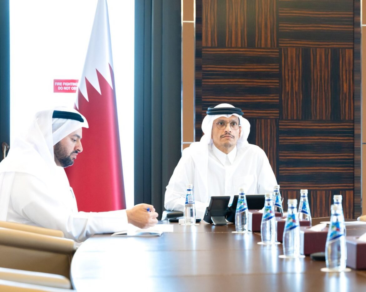 Two men in traditional Qatari clothing sit at a conference table with a Qatar flag in the background, during a formal meeting.