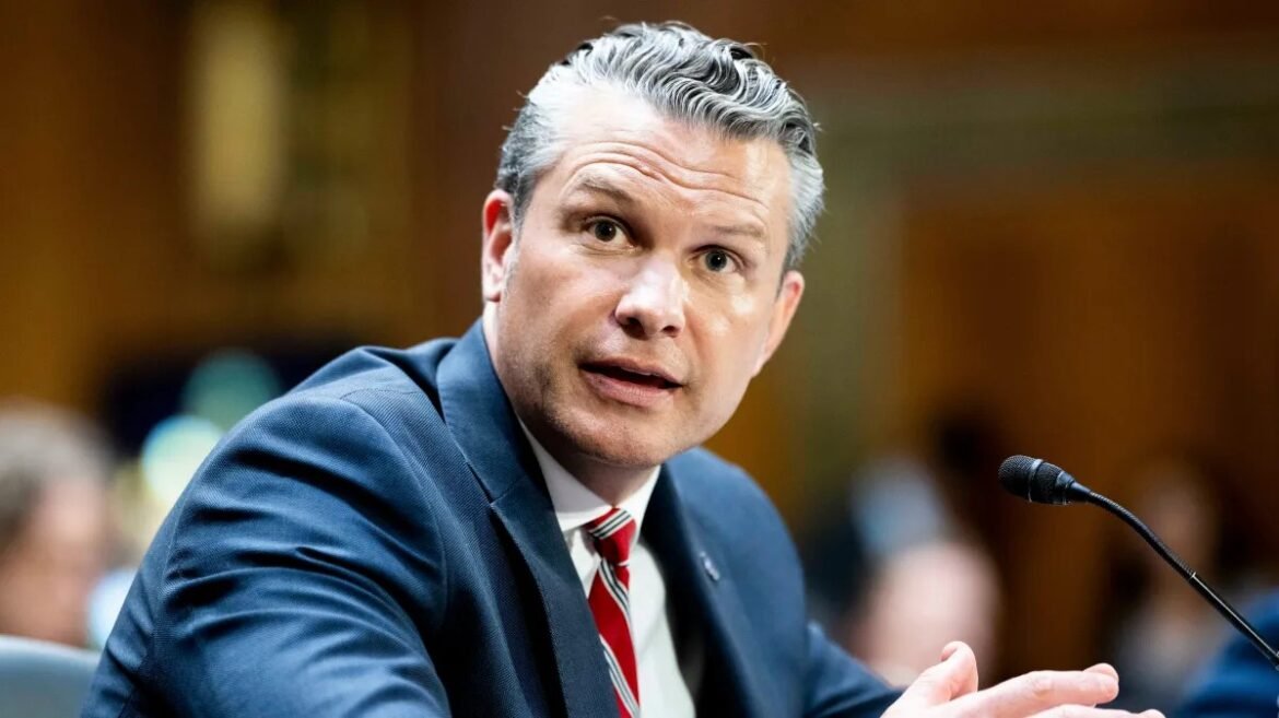 Man in a navy suit and red striped tie speaks into a microphone at a formal hearing.