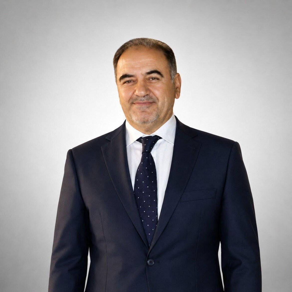 Smiling man in a navy suit, white shirt, and dark patterned tie against a light gray background (professional headshot).