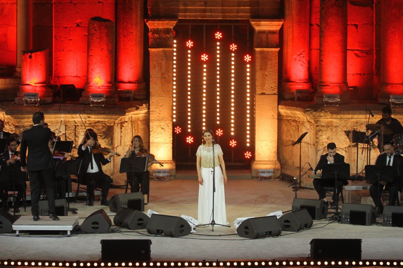 Female Singer in Cream Top and White Skirt Performs Center Stage with an Orchestra in a Red lit Ancient Ruin Backdrop  Aleumumnews