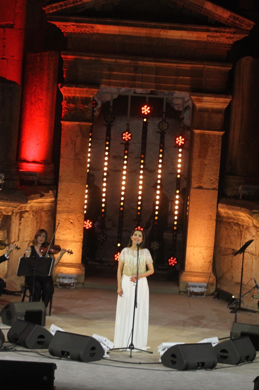 Female Singer in a Sparkly White Gown Performs on an Outdoor Stage at Night with a Violinist to the Left and Red lit Ancient Columns in the Background  Aleumumnews