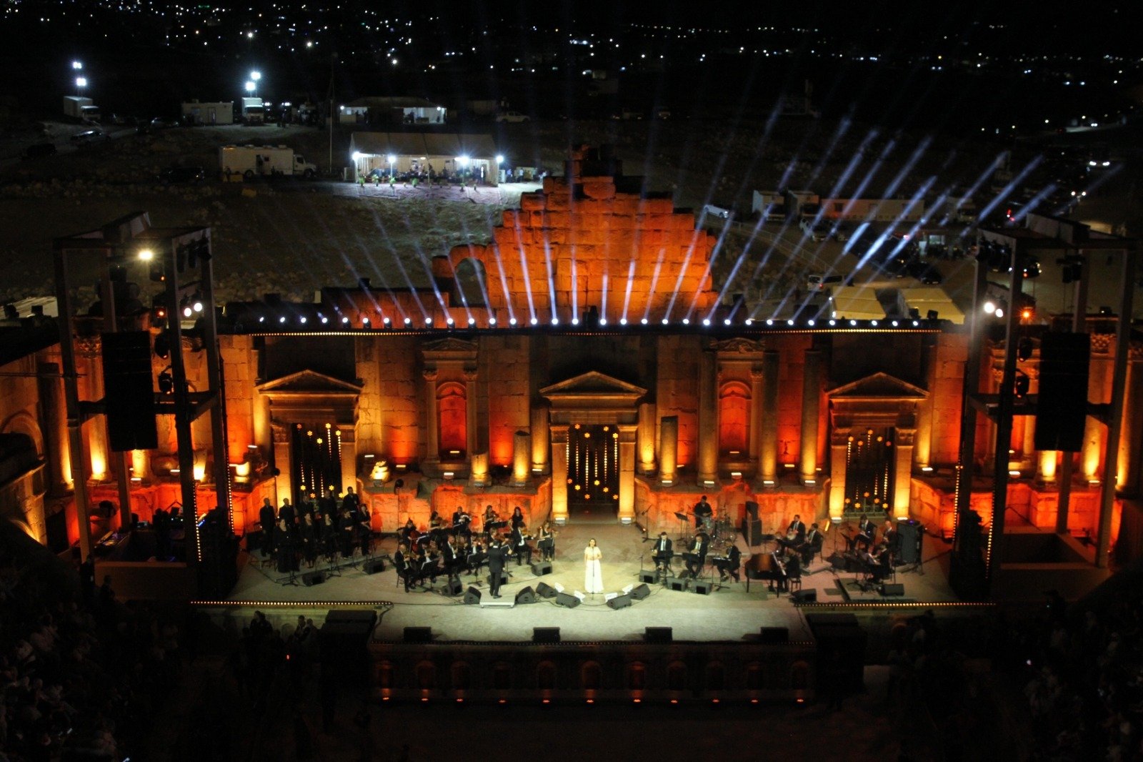 Nighttime Outdoor Concert on Ancient Ruin Stage with Orange Lights and a Full Orchestra Around a Solo Singer Center Stage  Aleumumnews