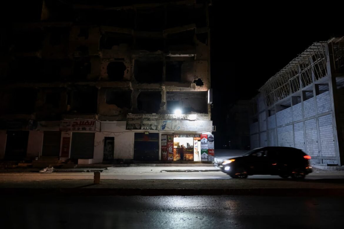 Nighttime urban street with a damaged multi-story building, a lit storefront at center, and a car passing by on the right.