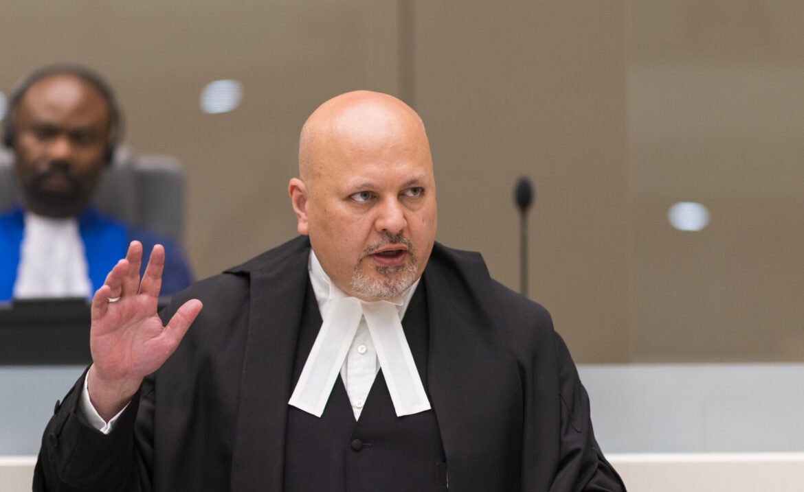 Bald judge in a black robe and white neckbands raises a hand while speaking in a courtroom; a colleague sits blurred in the background.