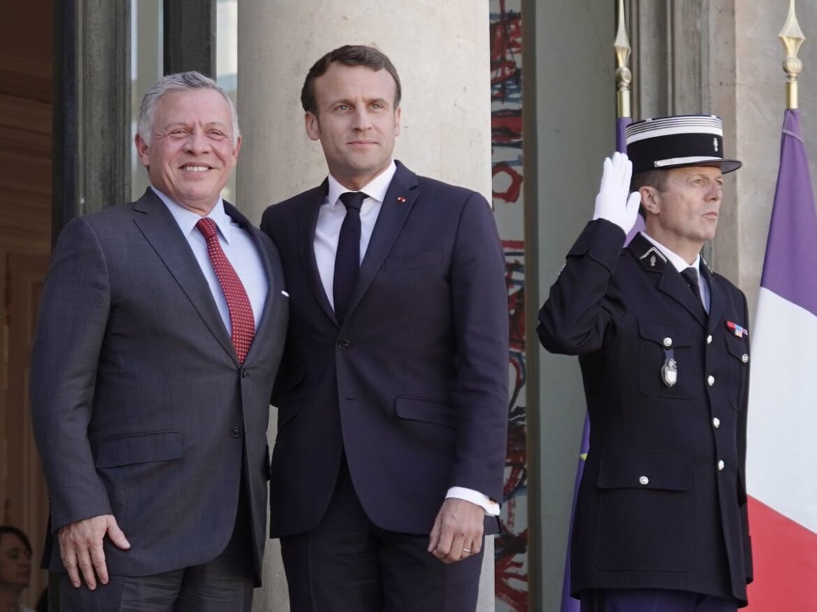 Two men in suits pose for a photo on a steps outside a building, with a uniformed officer saluting beside them and a flag in the background.
