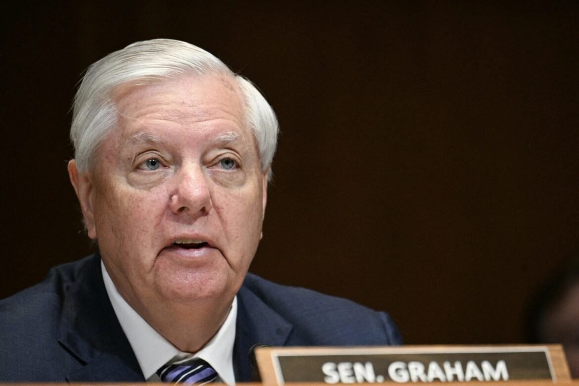 Close-up of an elderly man with white hair in a dark suit speaking at a desk during a formal hearing.
