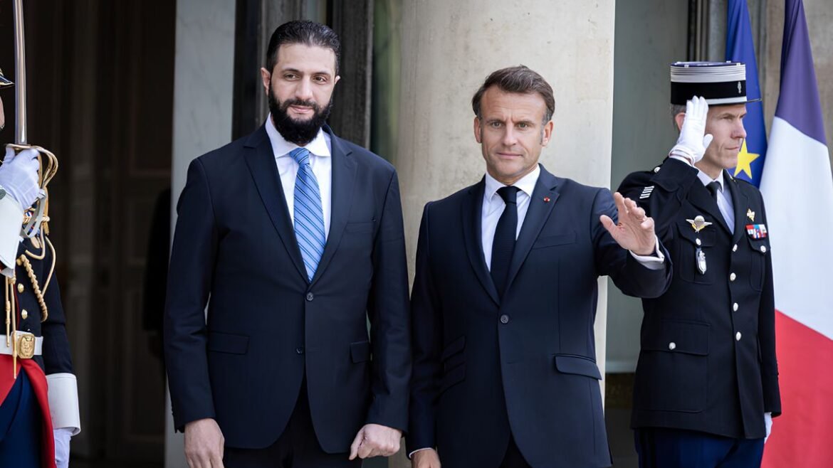 Two men in dark suits stand at a formal ceremony, a uniformed guard salutes beside flags in the background.